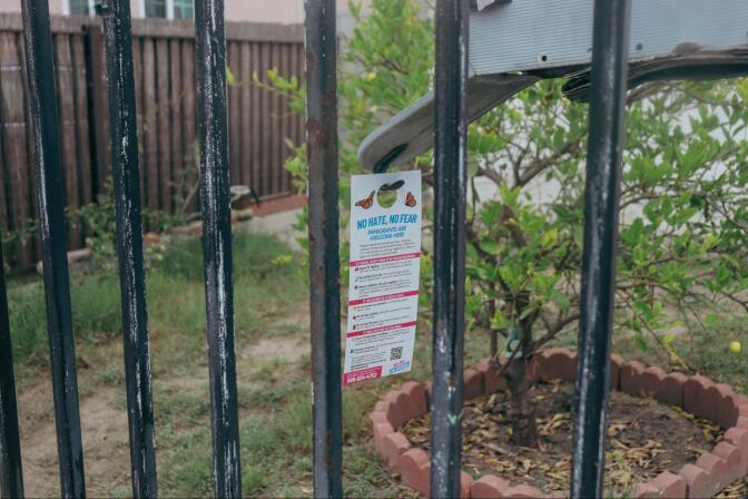 A small paper sign that reads "no hate no fear" hangs on a mailbox. The sign and mailbox are pictured through a black metal fence
