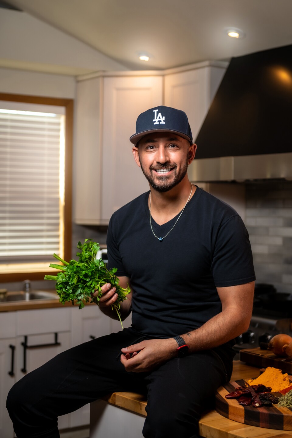 A photo of man with dark brown skin with a black mustache sits on kitchen island smiling holding a leafy green vegetable. 