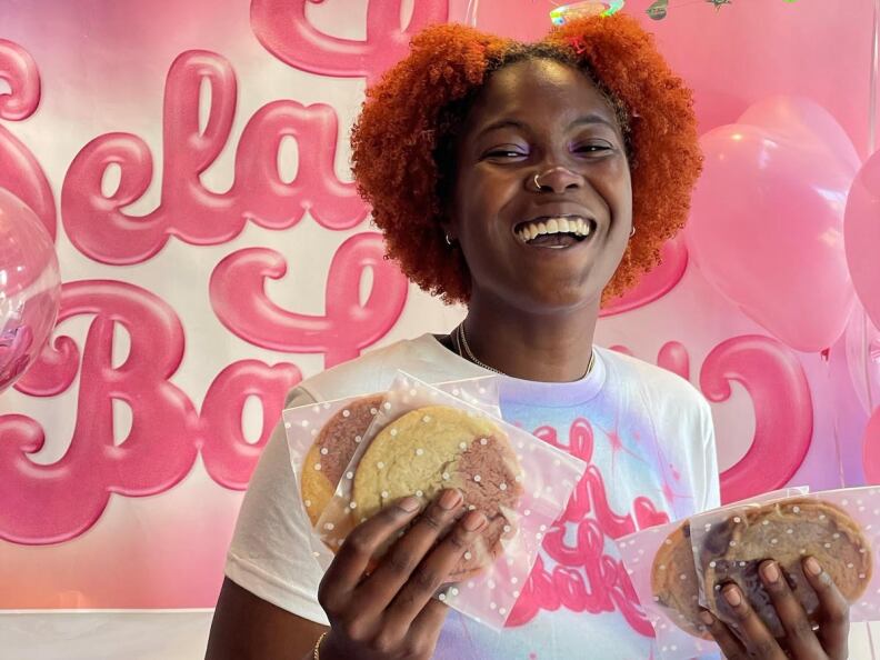 A woman smiling in front of a pink Selah Bakery sign holding cookies.