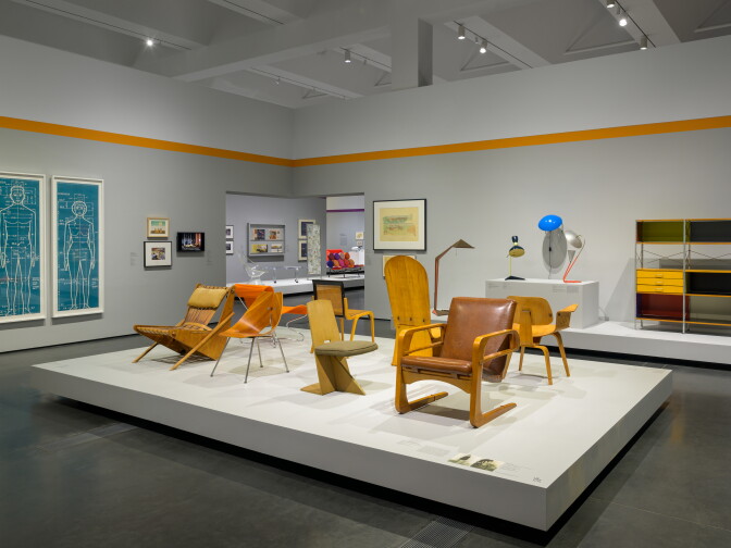Gallery view of mid-century modern chairs in blond wood and leather displayed on a white platform, with two blue anatomical blueprint-style drawings on the left wall and a colorful modular shelving unit and lamps on a pedestal at right.