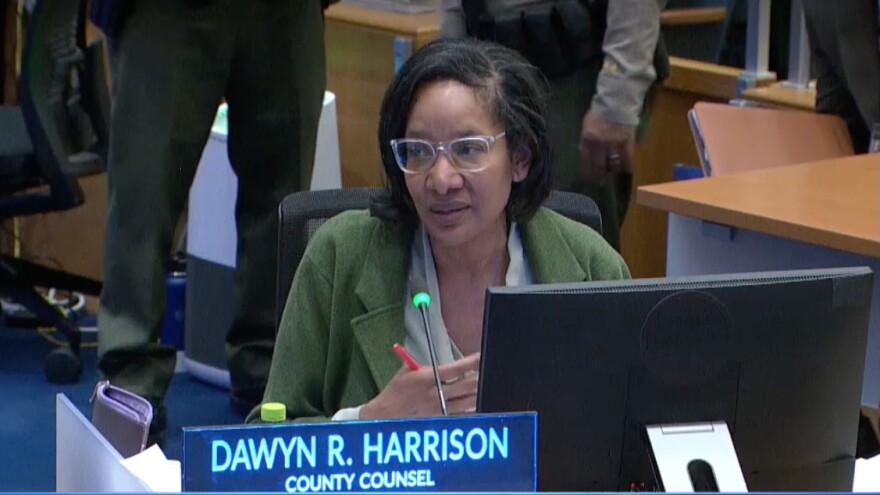 A Black woman wearing a green coat speaks into a microphone while holding a red pen at an official meeting, as a sign in front of her reads "Dawyn R. Harrison," "County Counsel."