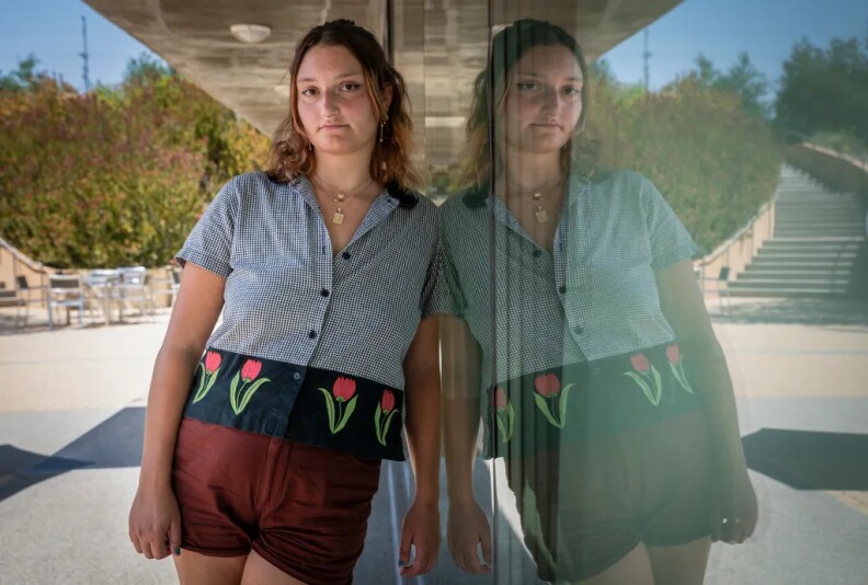 A portrait of a person leaning against a glass area so that they're reflection shows while they're looking at the camera.