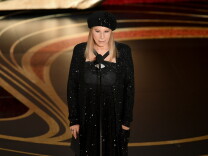 Barbra Streisand is shown onstage speaking into a microphone. She wears a long-sleeved black sequined gown and matching beret-style hat. 