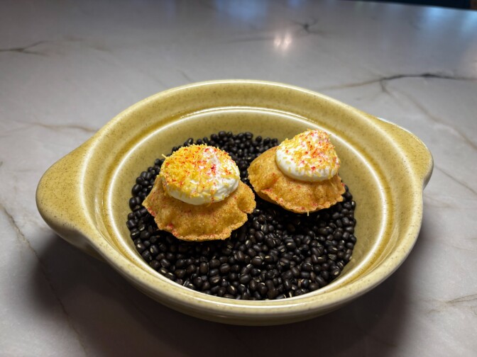 Two mini puris (crisp, hollow shells) lay on top of a bed of black bean shapes. They're crowned with whipped yogurt, and sprinkled with colorful savory dust. 