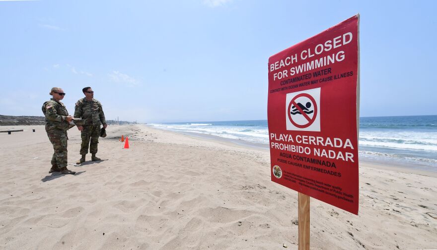 Two people in fatigues stand on the beach where a red sign warns: Beach Closed For Swimming. Contaminated Water.