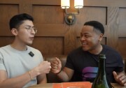 A young Latino man in an oatmeal colored t-shirt wearing glasses fist bumps a young Black man in a black t-shirt while sitting in a restaurant booth 