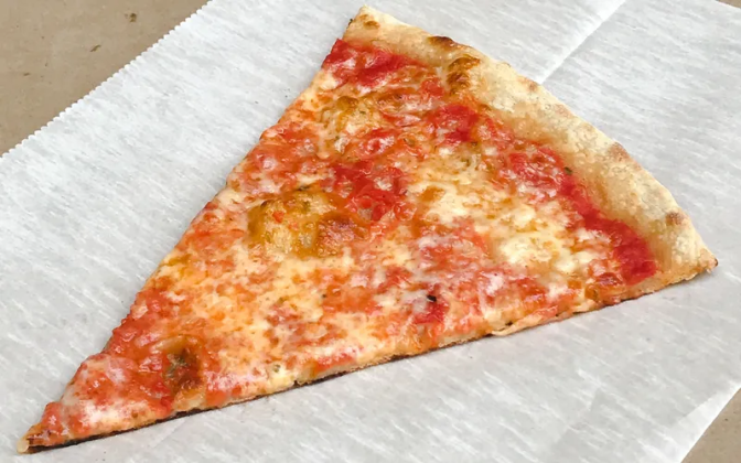A slice of cheese pizza is placed on a stark white piece of parchment paper on a beige surface. The triangular-cut slice contains melted white cheese on a bed of red tomato sauce, with a light brown crust at the top.