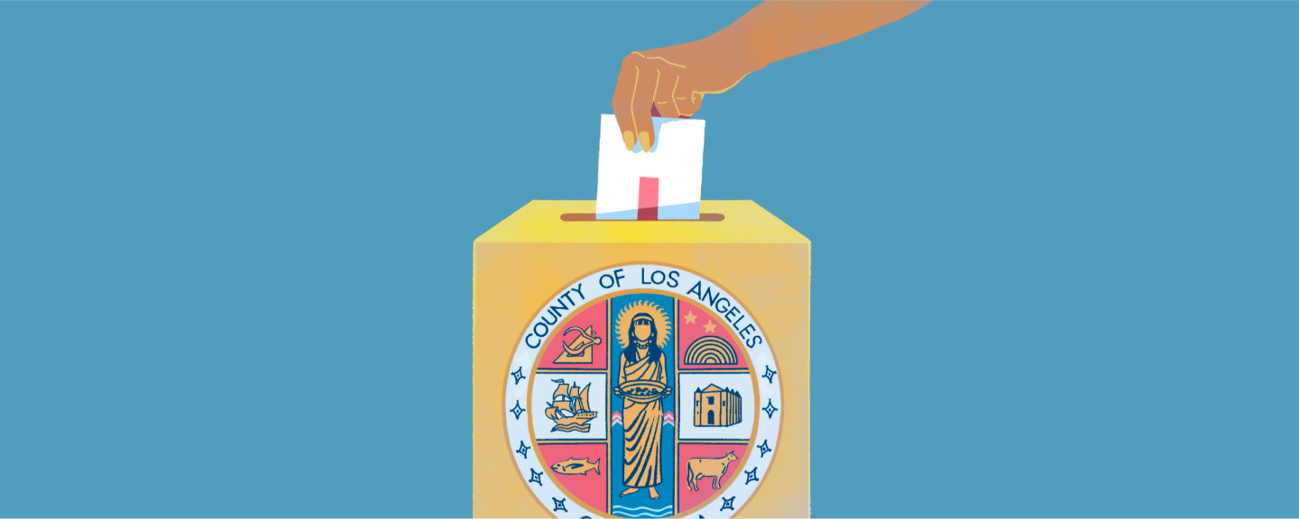 A person's hand inserts a ballot into a box marked with the L.A. County seal on the front of it.