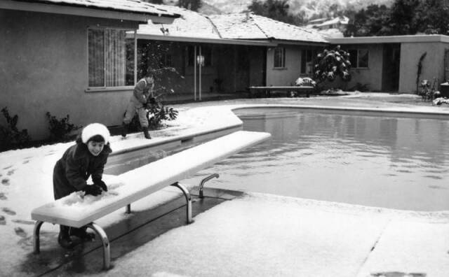 A black and white photo of two kids near a backyard pool scooping up piles of snow from the ground. One is scooping off the diving board. In the background are speckled mountains.