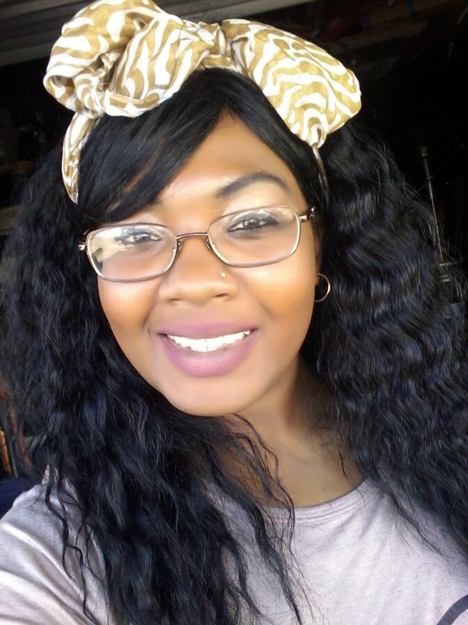A close-up image of a Black woman with long, wavy, black hair tied at the top with a bow smiles as she looks into the camera lens. She is wearing thin-rimmed glasses and pinkish lipstick. She also wears a white T-shirt.