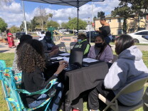 A group of women gathered around a table under a canopy. The table has a black tablecloth. Two of the women work at laptops, others wearing masks. Other women walk by in the background. They are sitting in chairs on grass, with a residential neighborhood behind.