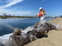 Plastic bags full of oil debris sit on the shore of a channel with a worker in white coveralls and an orange life preserver coils yellow rope.