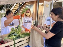 A female presenting person puts vegetables into a paper bag held by a female presenting person.