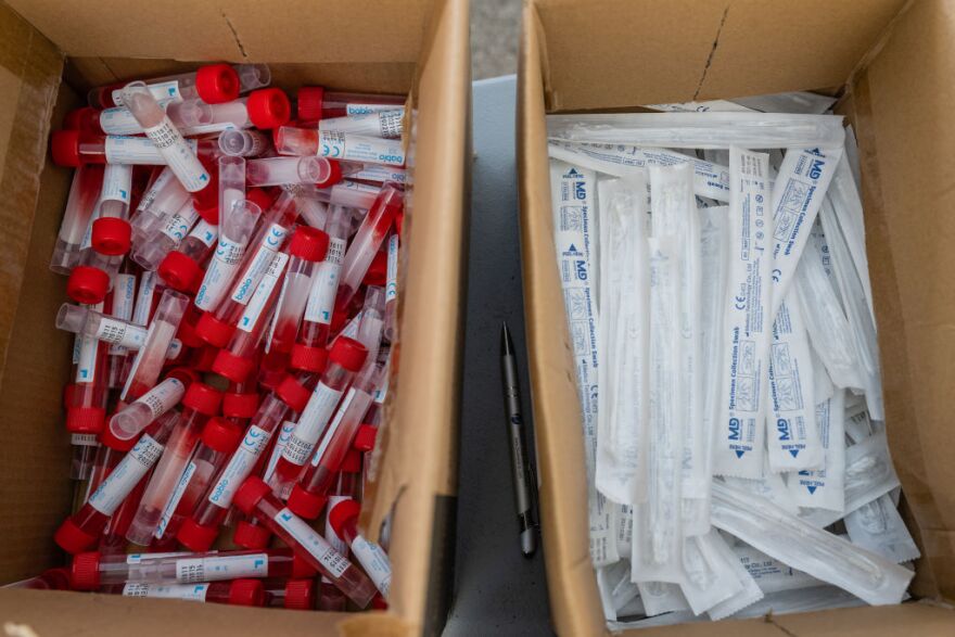 Two bins filled with COVID testing supplies sit side by side. On the left is a bin of red-capped vials containing testing fluid. On the right are unopened packages of nasal swabs.