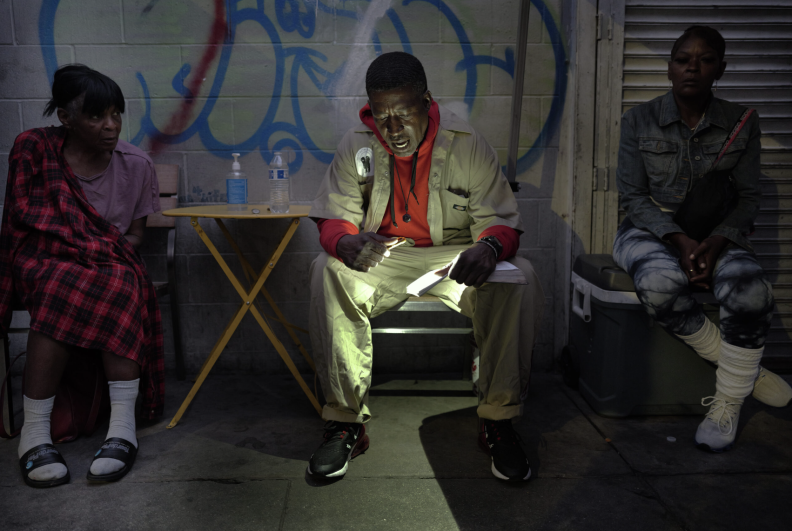 Three people with brown skin tone are sitting during the evening, facing toward the camera. One of them, a man, is in the middle using a light to read aloud from a book.