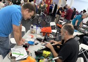 A room full of men and women of all ages. A white foldable table is set up closer to the camera, with two people seated behind it, including a man in blue jeans and a black t-shirt. Another man wearing a blue t-shirt and jeans is leaning over the table, which is covered in tools and electrical equipment.