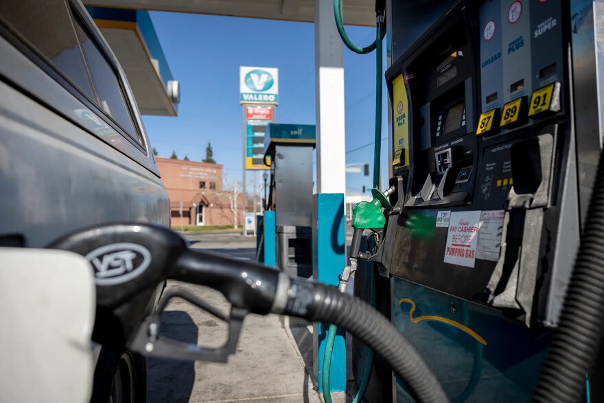 A close-up of a gasoline hose connected to a car. A Valero gas station sign can be seen in the distance.