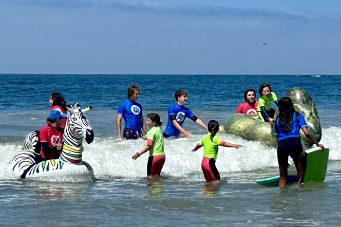 A small group of young adults and children stand in the shallow water wearing blue, neon green, and red shirts over wetsuits. One person is sitting on a unicorn-shaped raft and another is on a dinosaur-shaped raft. 