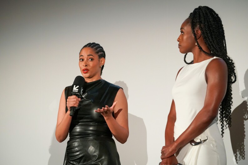 Two Black women stand beside one another. The woman on the left is wearing a black dress and is talking into a microphone. The woman on the right is wearing a white dress and is staring at the woman on the left.
