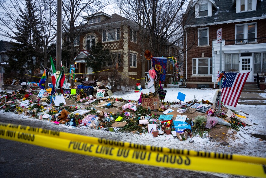 A memorial made of flowers, cards, flags, and signs on a pile of snow next to a sidewalk in front of homes covered in snow. The memorial is blocked off by police yellow caution tape.