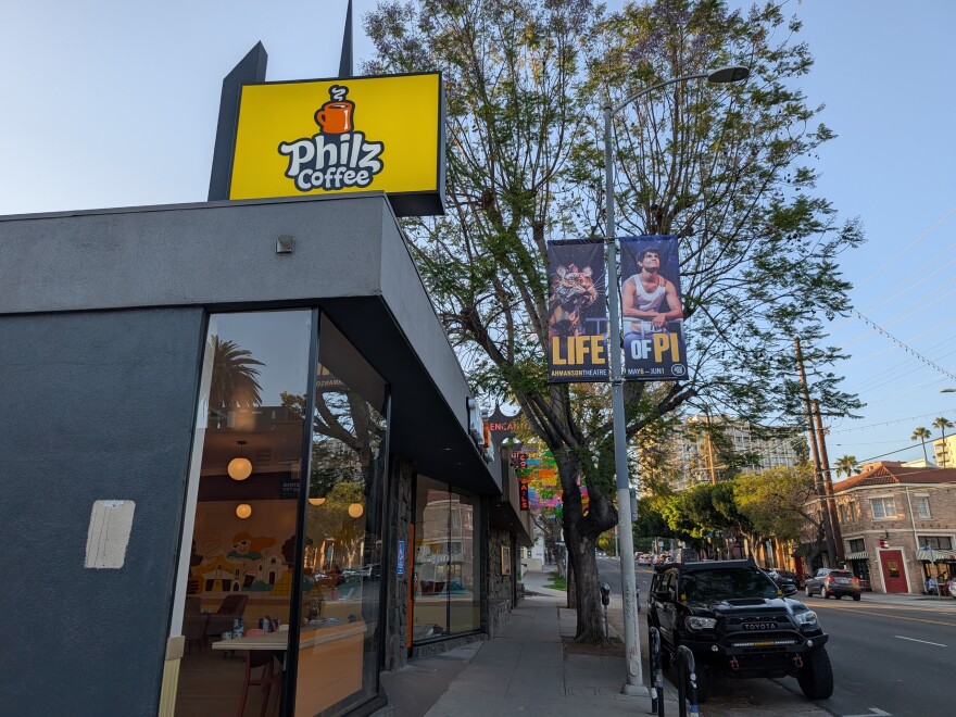The photo shows the exterior of a Philz Coffee shop, easily identifiable by a large yellow sign with the Philz Coffee logo. The shop features large windows, allowing a view inside where you can see hanging lights and tables.