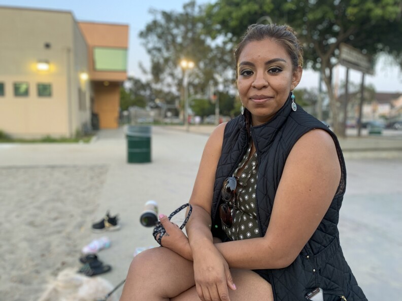 A woman with brown hair in a bun and a black vest sits at the park.