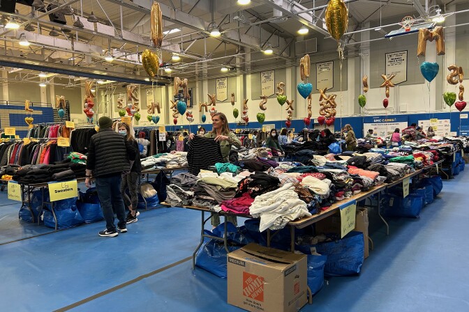 People peruse rows of tables laden with neatly-folded clothes. Mylar balloons are tied throughout the space. Some balloons are heart-shaped and some represent the letters "X," "M," "K," "S," and "L." They guide shoppers to tables with clothes that are their size. The tables are inside a basketball gym. 
