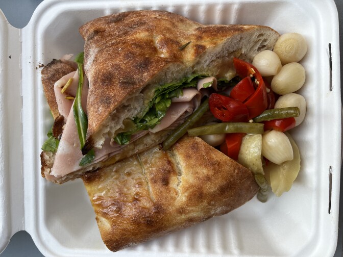 A sandwich with pale pink lunch meat coming out the side. The sandwich is cut in half, with one wedge stacked on top of the other, and is in a white plastic to-go container. Next to the sandwich is a small pile of vegetables containing small white onions, green beans, and red bell peppers. 