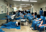 A room with white and green walls and bright lights is crowded with men whose faces are obscured in blue jail uniforms. Some are sleeping on the floor wrapped in blue blankets. Others are sleeping upright in chairs. 