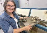 A woman with short brown hair, glasses and a jean jacket smiles into the camera while holding a small piece of concrete with oyster shells attached to it. She's standing in a room with several aquarium tanks.