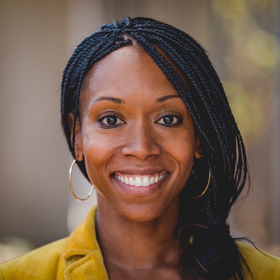 A Black woman with braids wears hoops and a yellow blazer.