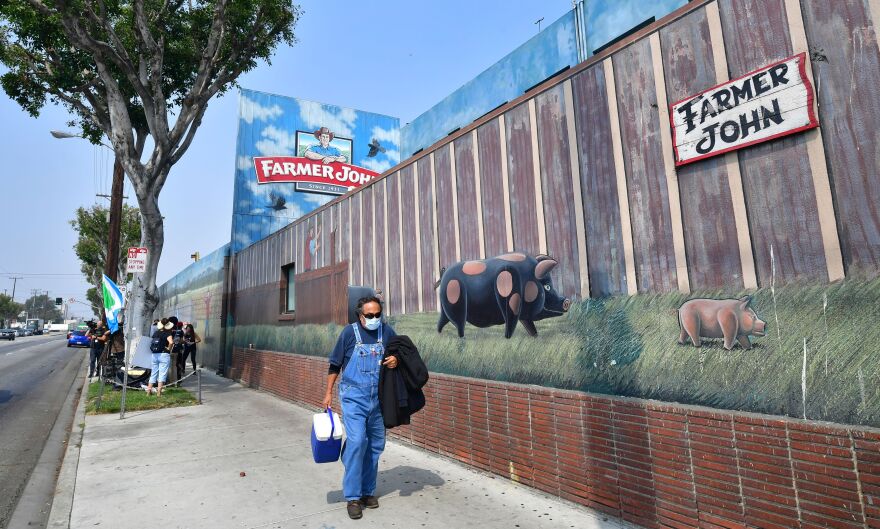 A person in overalls with mask on walks past the farmer john building on the sidewalk.