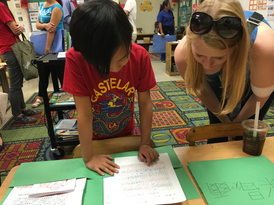 A young girl in a red shirt looks over a piece of paper. A woman with light skin leans over the table next to her.