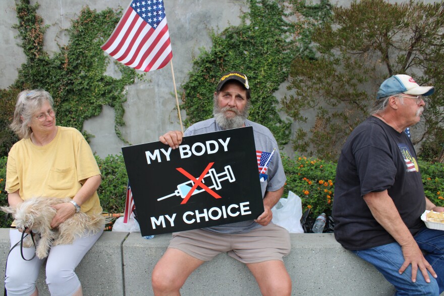 Three people sit on a ledge near plants. One person in the middle is holding up a black sign that says "my body my choice" and an American flag. On the middle of the sign is a vaccination needle with an x through it.