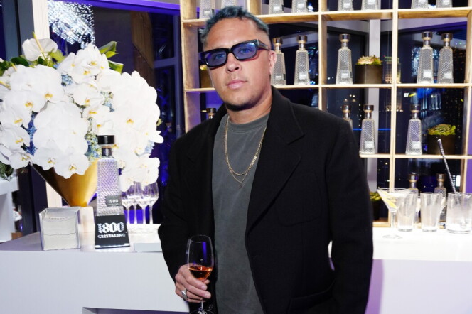 A Brown man wearing a black jacket and pants holding a beverage poses for the camera in front of a well-lit bar.