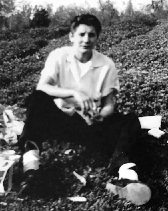 A black and white view of a Mexican American woman, with a light colored button up, sitting outside on the ground by different plants. She's sitting wtih her knees pulled up while resting her elbows on them.