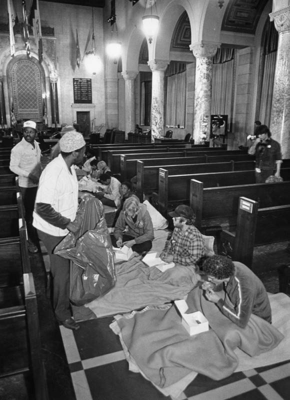 A black and white view of a row of people, with different skin tones and genders, sit on the ground with blankets and food in between the council chamber's benches indoors. There are people walking around them carying bags.