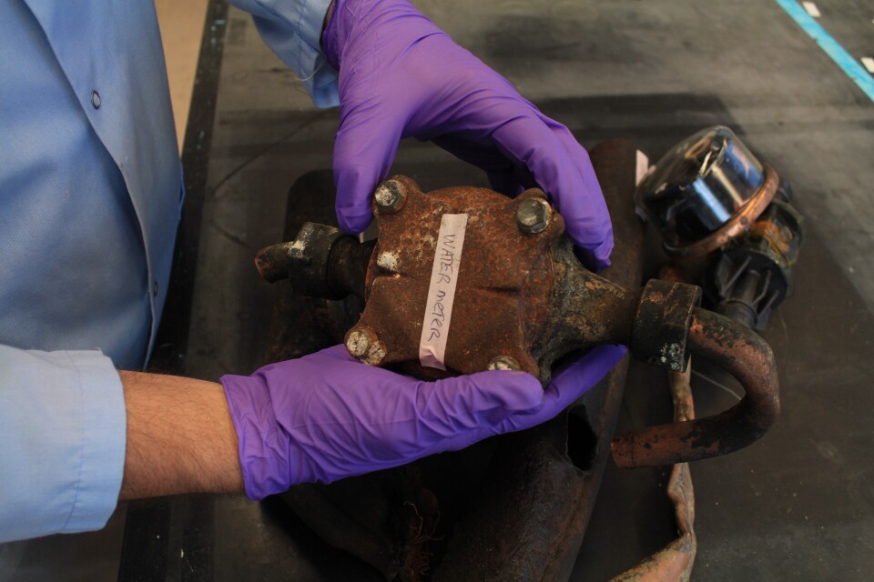 Closeup of a person wearing a blue lab coat and purple plastc gloves holding a rusted piece of equipment labeled "water meter."