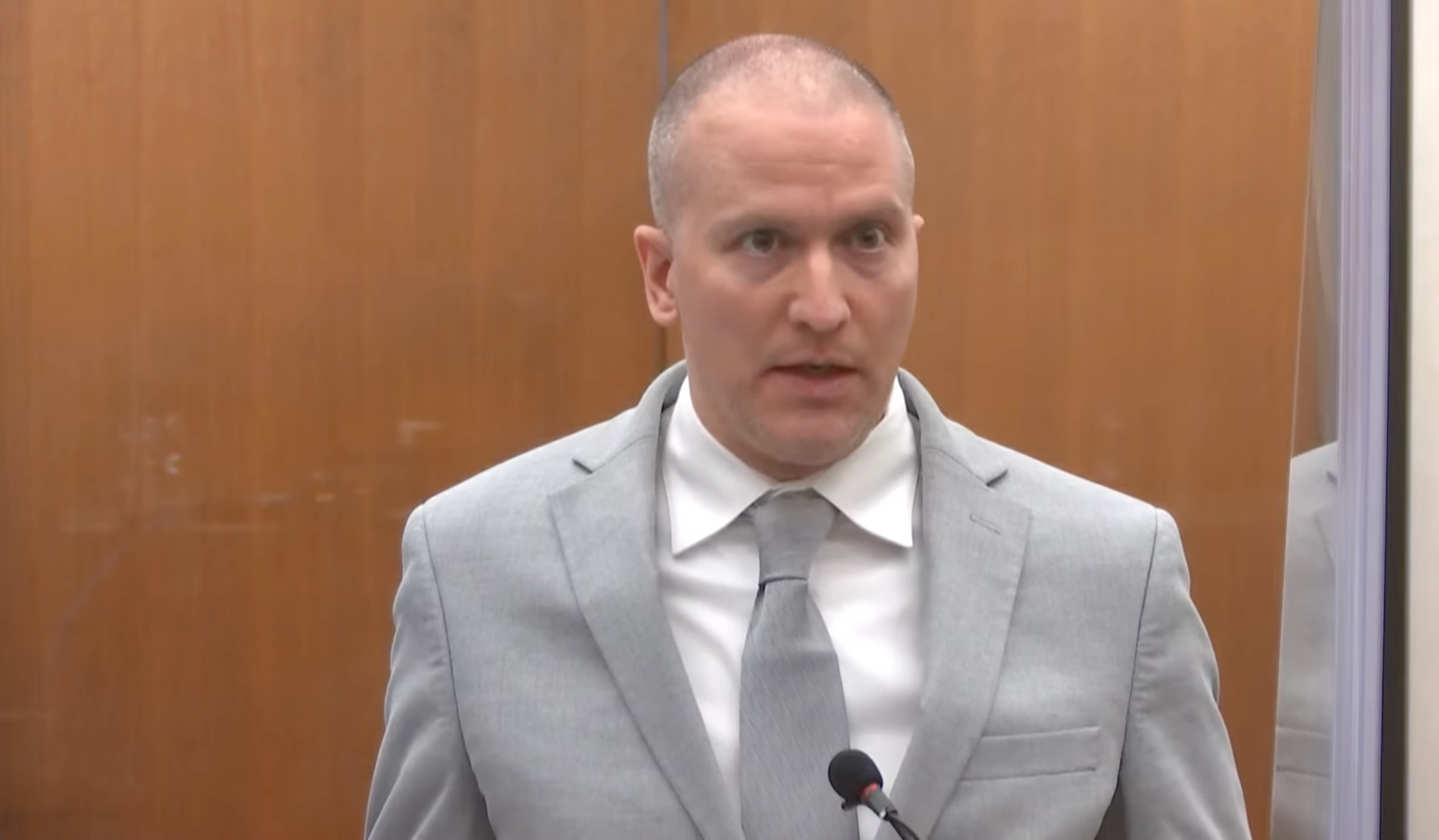 Former police officer Derek Chauvin is shown seated in a Minneapolis courtroom, wearing a light gray suit with a dark blue shirt and dark blue tie and a yellow lanyard around his neck, looking up toward the bench.