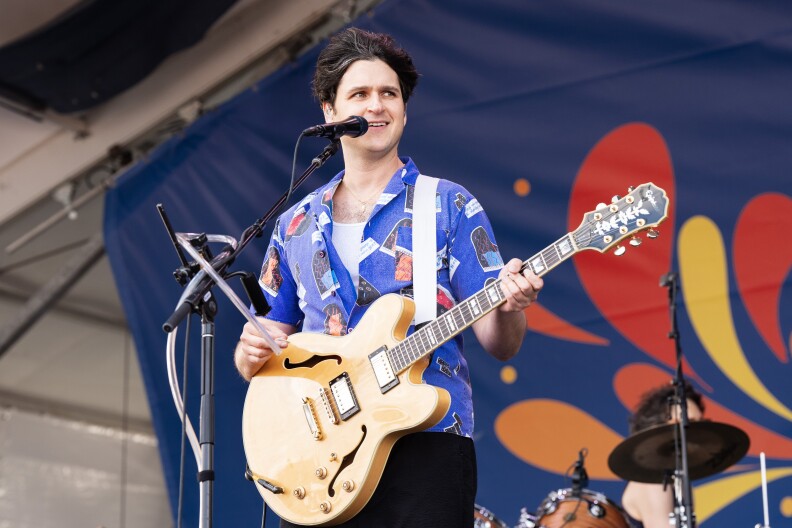 Exra Keonig performs on stage, playing a tan electric guitar and speaking into a micrphone. Behind him is a drummer and a blue, red, orange, and yellow background.