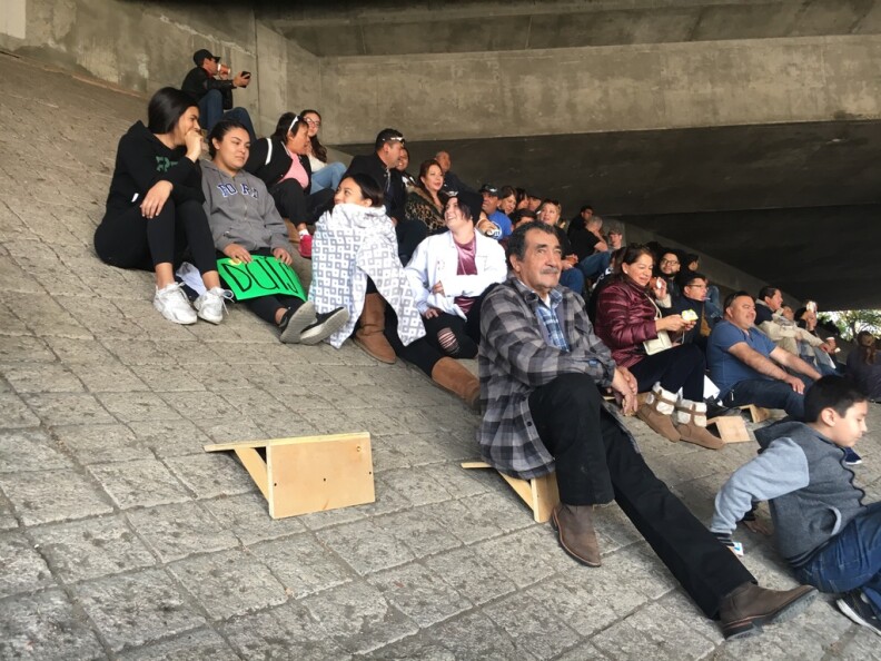People sit on small wooden seats that help them perch on a sloped wall under a freeway overpass
