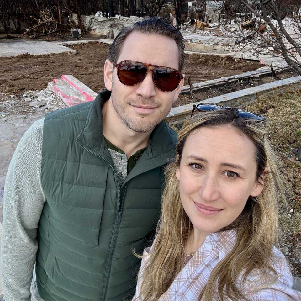 A younger middle aged couple with light skin tone takes a selfie. The woman on the right has long blond hair, and her husband, wears sunglasses and a green vest. Behind them is the rubble of their burned home. 