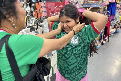 A young, female presenting person holds her arms up and wears a green sports jersey. Her mother holds her cheeks in a caring way.