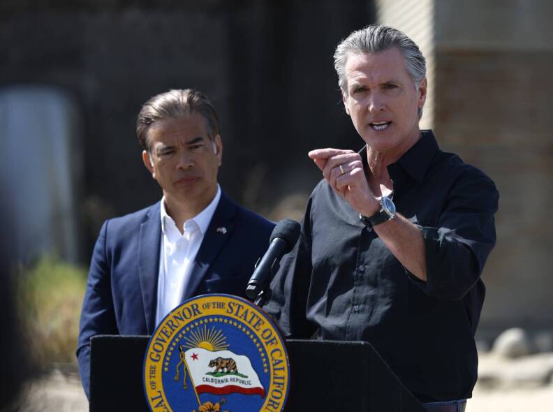 Gov. Gavin Newsom, a man with light skin tone wearing a black shirt with the sleeves rolled up, speaks into a microphone at a podium while raising his left hand. Attorney General Rob Bonta, a man with medium skin tone wearing a blue suit and unbuttoned shirt, listens to the the right behind him.