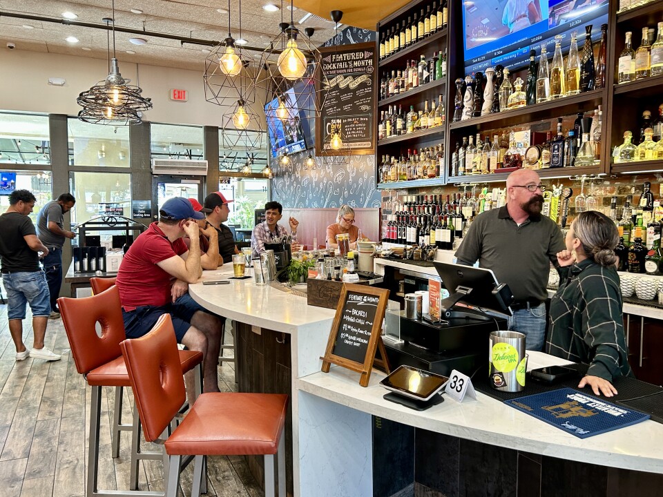 Four men and one woman sit at a brightly lit semi-circle bar. Alcohol lines the shelves behind the bar and two workers discuss something behind the cash register. 