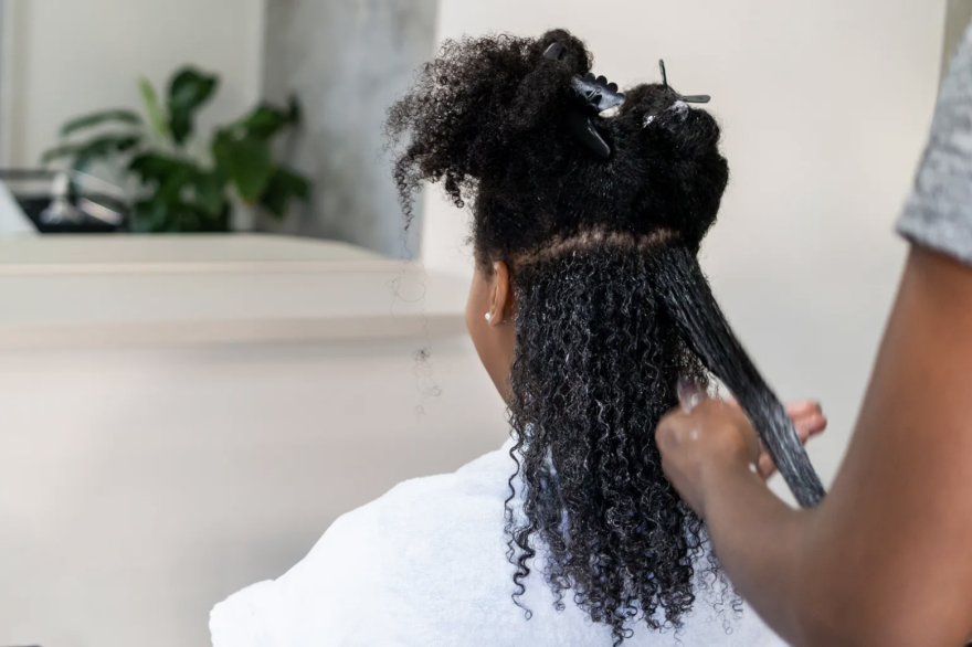 A Black woman sits in a chair facing away from the camera, getting her hair straightened.
