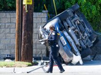 A uniformed police officer walks past a silver sedan that is on it's side, resting against a hollow block fence. There is a wooden electrical pole to the left of the vehicle.