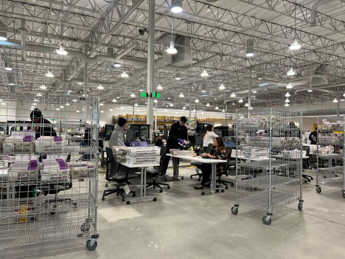 Election workers in a large warehouse with fluorescent lights suspended from ceiling girders sit at tables with boxes filled with votes for processing