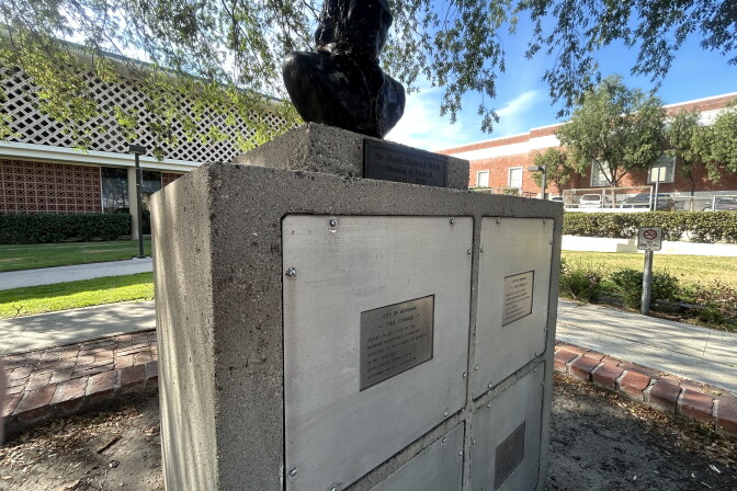 A cement monument with four square compartments topped by a black bust of a man. The monument is outside on a sunny day between two buildings and patches of grass.