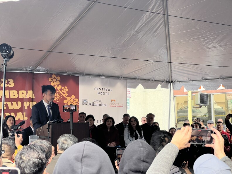 A crowd under a tent looks up at a a young Asian man with a mustache and beard who stands at a podium, speaking